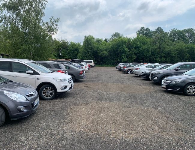 Aparcamiento con vistas – El terreno es de grava y está rodeado de árboles. Muchos coches están en fila a ambos lados.