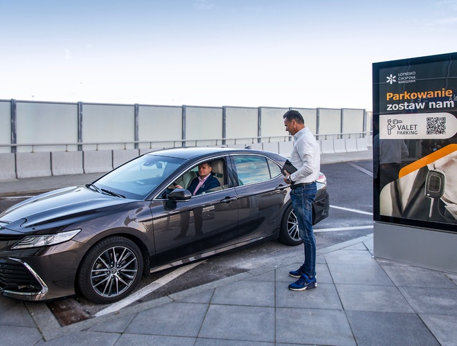 Valet Parking - Aeropuerto Chopin - El conductor en el coche conversa con el encargado del estacionamiento. Al lado, un letrero con el logo de Valet Parking. Superficie de losas de pavimento.