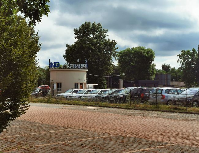 Parking Tani i Bezpieczny PREMIUM - Obiekt jest ogrodzony siatką. Na parkingu stoi wiele samochodów. Widać budynek z napisem.