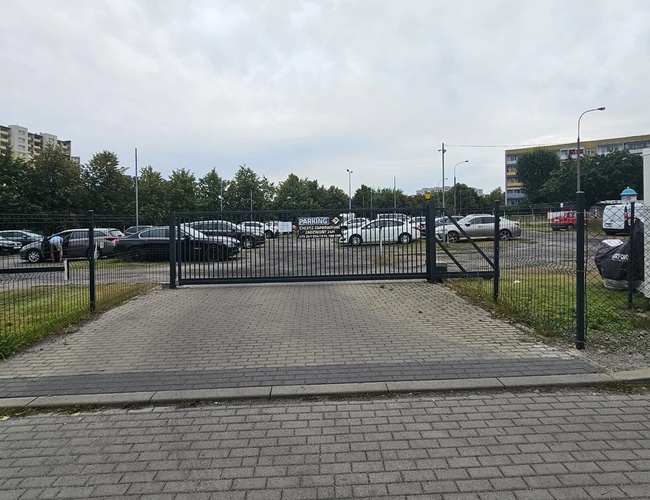 Odlotowy Parking BZG - Parking jest ogrodzony metalowym płotem. W tle stoją zaparkowane samochody.