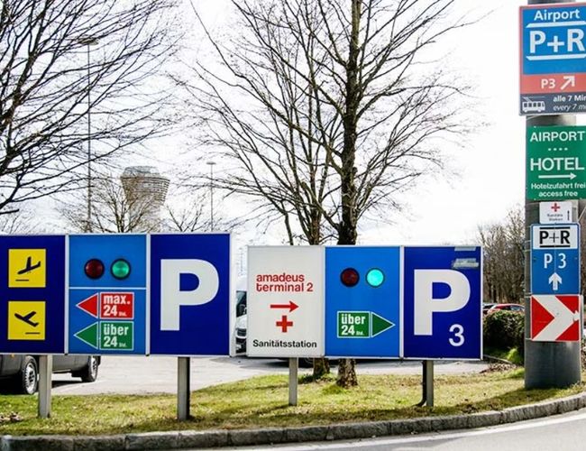 Zespół tablic kierunkowych prowadzących do różnych stref parkingowych. Na znakach znajdują się oznaczenia parkingów oraz kierunki dojazdu.