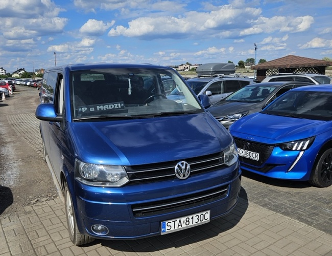 Aparcamiento en Madzi – están estacionados coches particulares. El terreno no está claramente marcado. Cielo despejado al fondo.