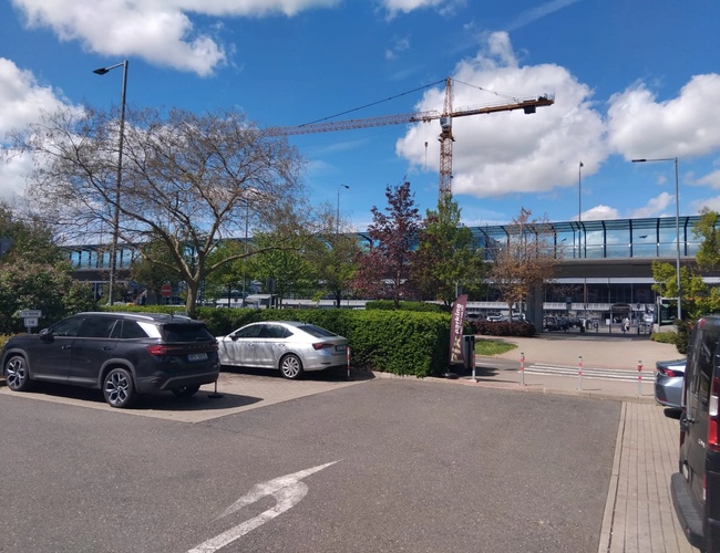 PRG Parking - Na parkovisku stoja dve autá. Vidieť žeriav a zelené rastliny. V pozadí je moderná budova.