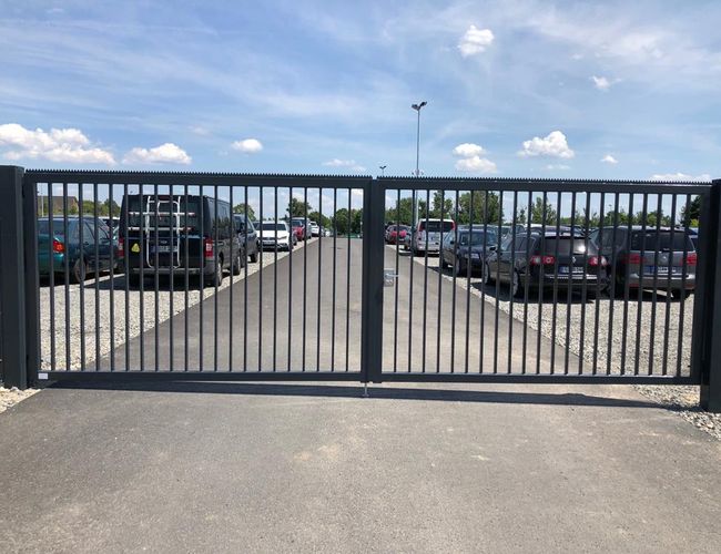 A parkolóhelyed – A terület kerítéssel körülvett és kiépített. Mindkét oldalán parkolt autók állnak. Háttérben fák láthatók.