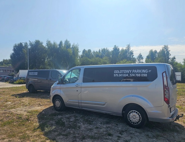 Odletové Parkovisko Mado - Dve dodávky stoja na tráve. Na pozadí sú stromy a budova.