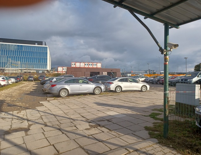 Maxi Parking – Se pueden ver varios coches aparcados. El área está parcialmente cercada. También se puede ver un techado y una cámara.