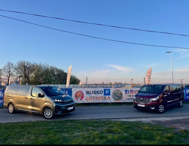 Parking Blisko Lotniska – Na kostce stoją dwa busy. W tle ląduje samolot. Widoczne są drzewa i zachmurzone niebo.