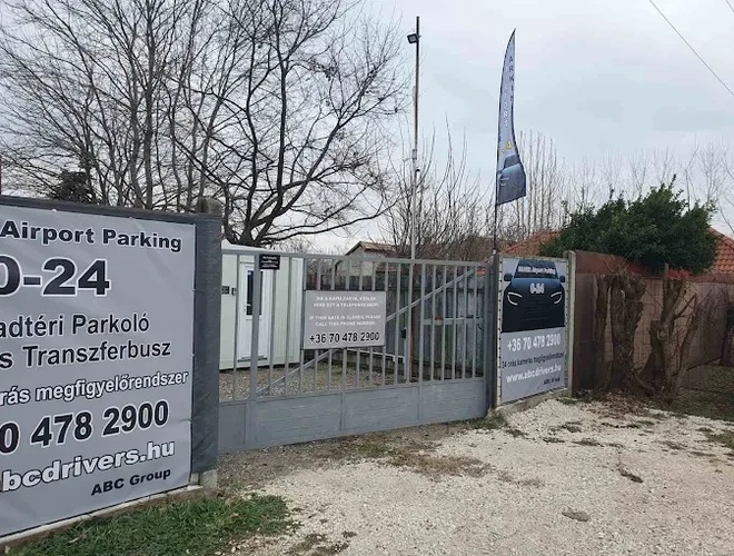 Widoczna brama wjazdowa na parking z banerami reklamowymi. Teren ogrodzony, dookoła drzewa.