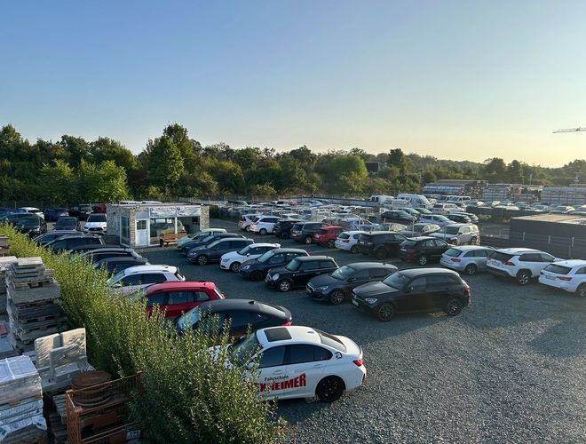 Shuttle Parking Vezető - A parkoló tele van autókkal, amelyek kavicsos talajon állnak. Mellette található egy kis üveg-kő ügyfélszolgálat. Körülötte zöld növényzet van.