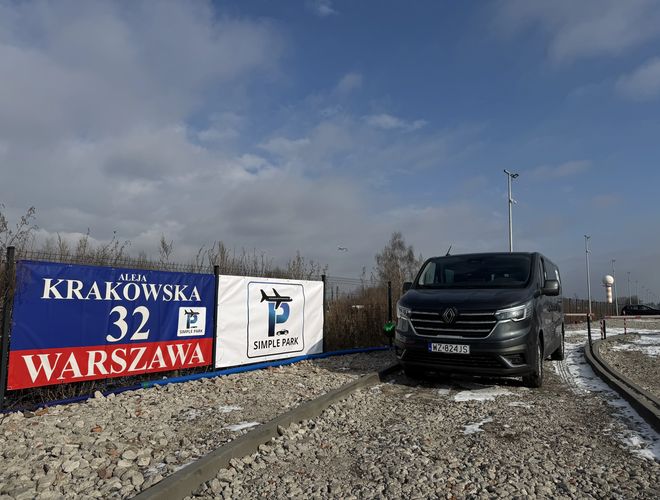 Parking Simple Park – na żwirowym placu stoi szary samochód. Widoczny jest także baner z adresem w tle. Po lewej stronie widać ogrodzenie.