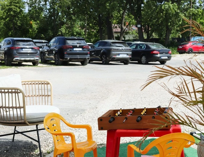 Najlepšia parkovacia séria A – Stoja tam autá v tieni stromov. Vpredu sú stoličky a stôl na hranie. Na pozadí je vidieť zeleň.