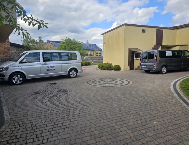 Estacionamiento en Krótka - Área pavimentada con adoquines. Dos furgonetas están al lado del edificio. Al lado, arbustos y un entorno ordenado.