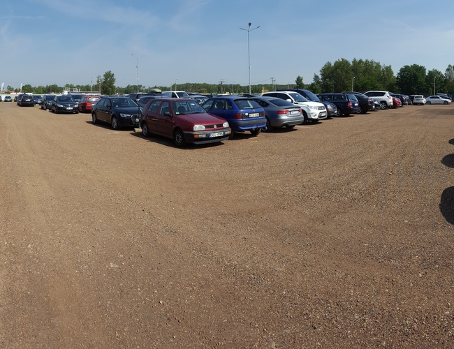 AAP-66 – Parking jest żwirowy i otwarty. Widać wiele zaparkowanych samochodów. Otacza go płot i latarnie.