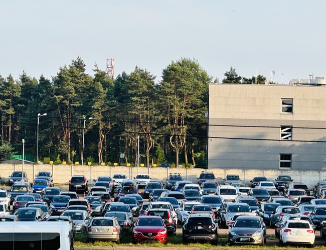 Comienza con nosotros - En el aparcamiento hay muchos coches. Al lado hay un bosque y un edificio. El área está cercada.