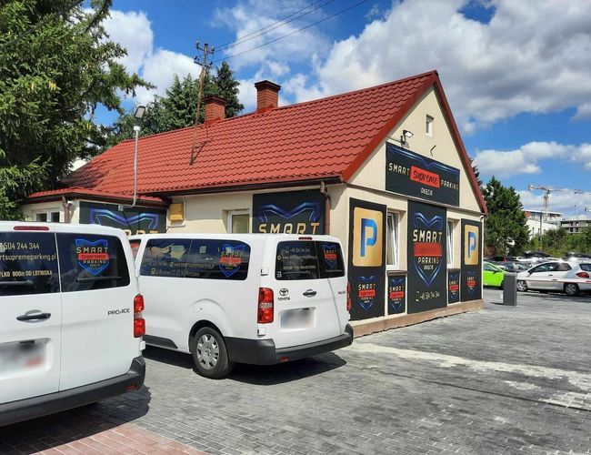 Smart Supreme Parking - Budynek z czerwonym dachem i szyldy. Przed nim zaparkowane są białe pojazdy. Obok widać także inne samochody.