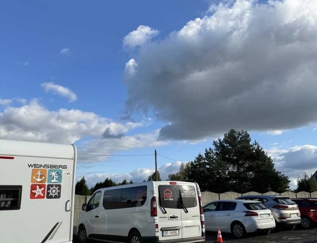 Aparcamiento Wings – En el aparcamiento hay coches y una caravana. El terreno está cercado y rodeado de árboles. Sobre el aparcamiento se puede ver el cielo azul y las nubes.