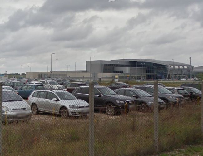 Oficjalny Parking P6 – Teren jest ogrodzony siatką. Widać wiele zaparkowanych samochodów. W tle stoi duży budynek.
