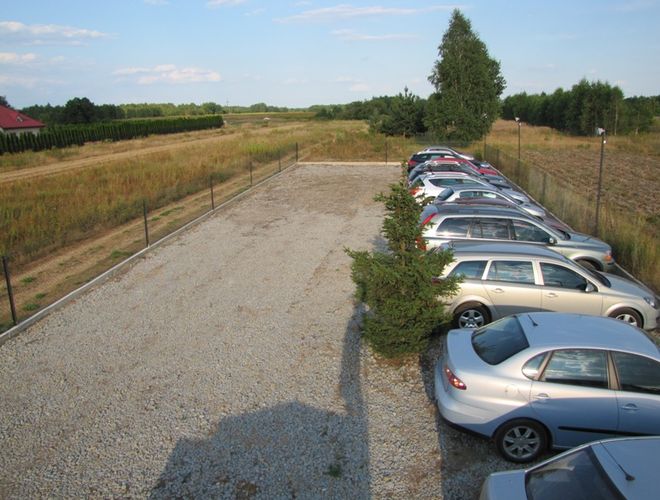 Parking Auto Park - Rząd aut stoi na żwirowym placu. Parking jest ogrodzony i otoczony drzewami.
