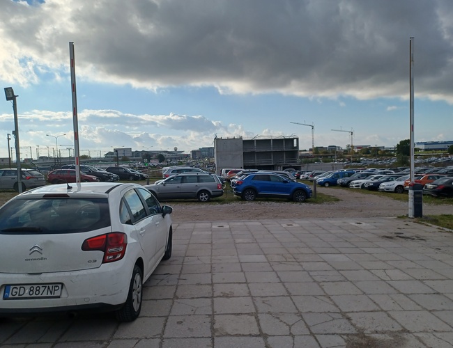 Parking Goliat – na placu znajdują się samochody osobowe. Teren jest częściowo utwardzony płytami. W tle widać dźwigi budowlane.