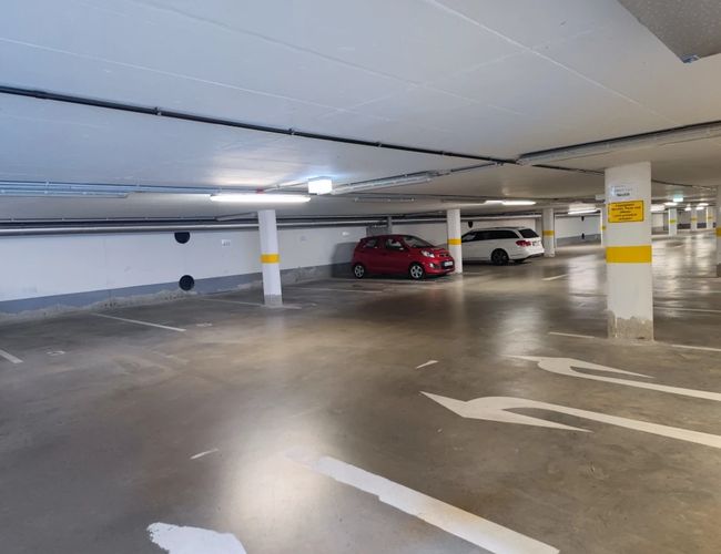 Parking FrankfurtValet24 - Podziemny parking z niskim sufitem. Dwa zaparkowane samochody. Jasne oświetlenie zapewnia dobrą widoczność.
