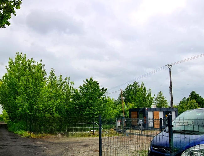 Krizpit Parking 24 – ohradené parkovisko s mrežou a jedným závorou. Na pozadí je vidieť stromy a sivú oblohu. Na dvore stojí modré auto.