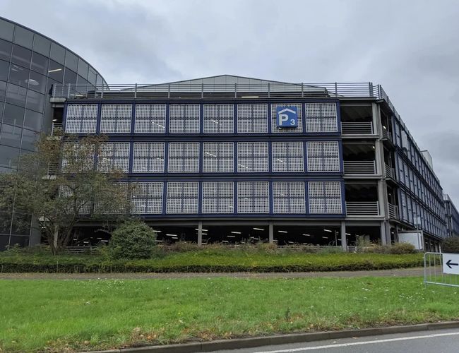 Zewnętrzny widok na nowoczesny parking wielopoziomowy z przeszkloną fasadą, położony w pobliżu terminala lotniska i głównych dróg dojazdowych.