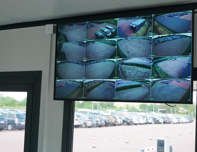 VIP Garáže MEGA Parkovisko – Monitor s obrazom z kamier. Vidno rôzne zábery parkoviska. V pozadí stoja zaparkované autá.
