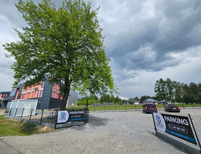 ParkZone - Parking jest ogrodzony i oświetlony. Stoją tam dwa samochody. Obok niego znajduje się duże drzewo.
