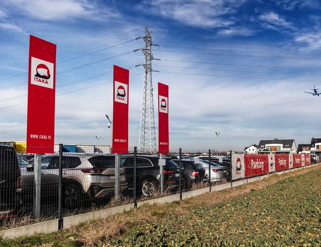 Parking Okęcie Południe – wiele zaparkowanych samochodów. Teren jest ogrodzony wysokim płotem. Nad parkingiem leci samolot.
