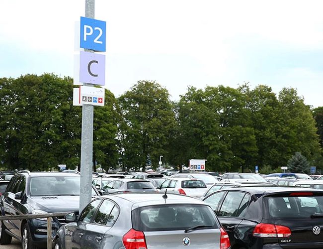 Parking z zaparkowanymi samochodami. Na pierwszym planie znajduje się słupek z oznaczeniem sektora parkingowego „P2 C”, a w tle widoczne są drzewa.