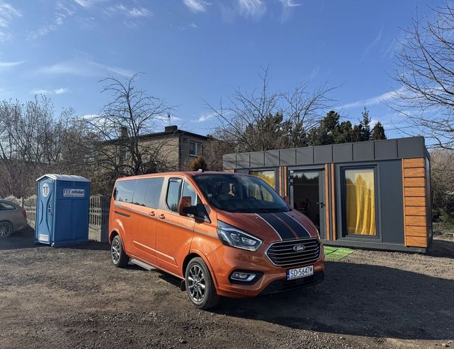 Parking Wings - En el área hay un autobús naranja. Junto a él hay un inodoro portátil azul y un edificio con cortinas amarillas. En el cielo hay nubes suaves.