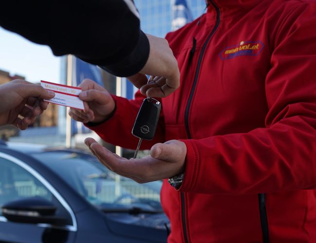 Parking Mein Valet Outdoor - Osoba przekazuje kluczyki i dokument pracownikowi w czerwonej kurtce. W tle stoi zaparkowany samochód.