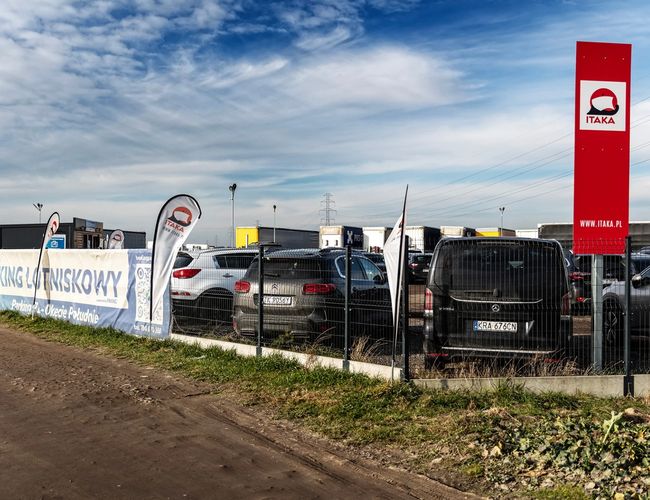 Parking Okęcie Południe - Ogrodzony teren z samochodami i banerami reklamowymi. Czysta, asfaltowa nawierzchnia. Teren oświetlony latarniami.