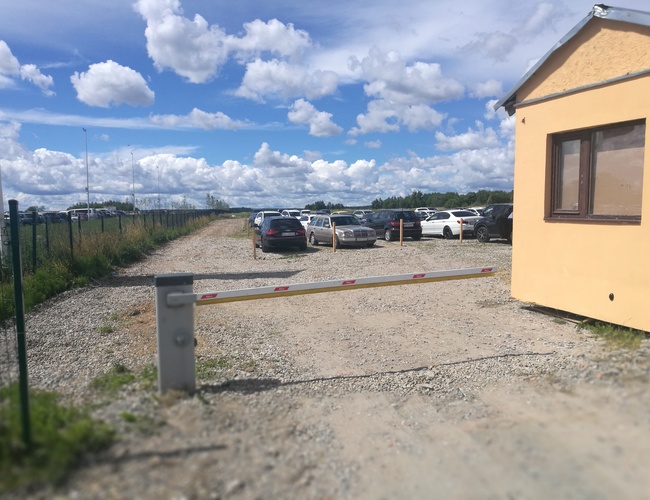 Parking P8 – Widoczny szutrowy plac z samochodami. Ogrodzenie biegnie wzdłuż lewej strony. Szlaban przy wjeździe.