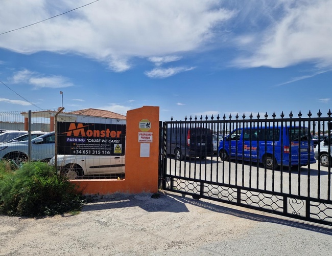 Monster Parking - Oplotené parkovisko s kovovou bránou. Sú na ňom zaparkované autá. Vedľa brány visí informačná tabuľa.