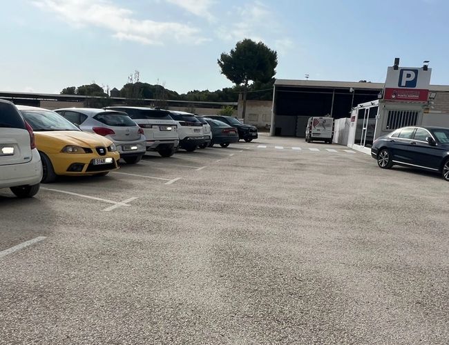 Parking Punta Marina – Na parkingu stoi wiele samochodów. W tle widoczny jest budynek z bramą wjazdową. Pogoda jest słoneczna.