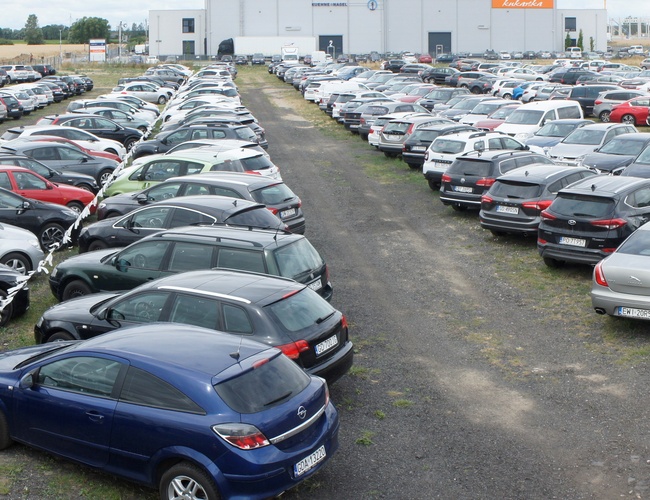 Parking Lotniskowy 24h – Duży parking zapełniony autami. Teren jest utwardzony i oświetlony. Parking otacza niski płot.