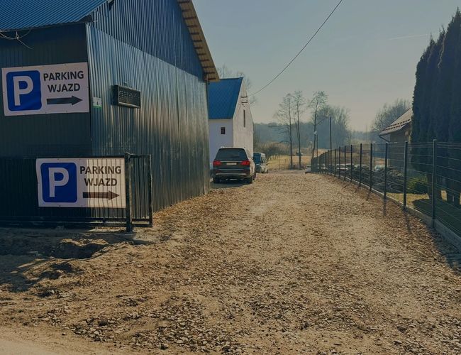 Parking Amber – Żwirowy dojazd między blaszanym budynkiem a siatką. Znak wskazuje wjazd na parking. Po lewej stronie widać zaparkowany samochód.