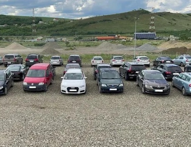 Parking pełen samochodów ustawionych na żwirowym placu, w tle zielone wzgórza i niebo z chmurami.