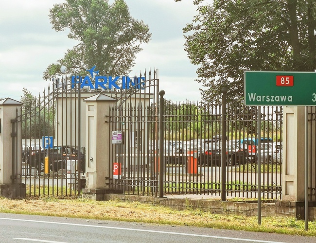 Dostupné a bezpečné parkovisko PREMIUM - Kovový plôt a brána s nápisom. Na pozadí zaparkované autá. Vpravo tabuľa s smerom do Varšavy.