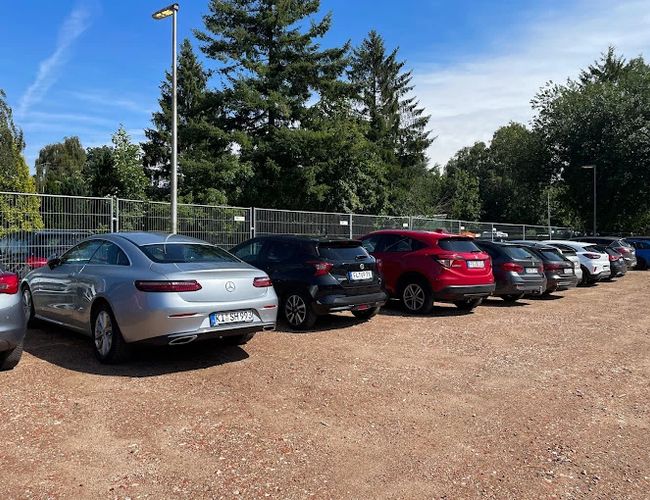 Parking Park In Fly Out – Na parkingu stoją samochody. Teren jest ogrodzony i oświetlony. W tle widoczne są drzewa.