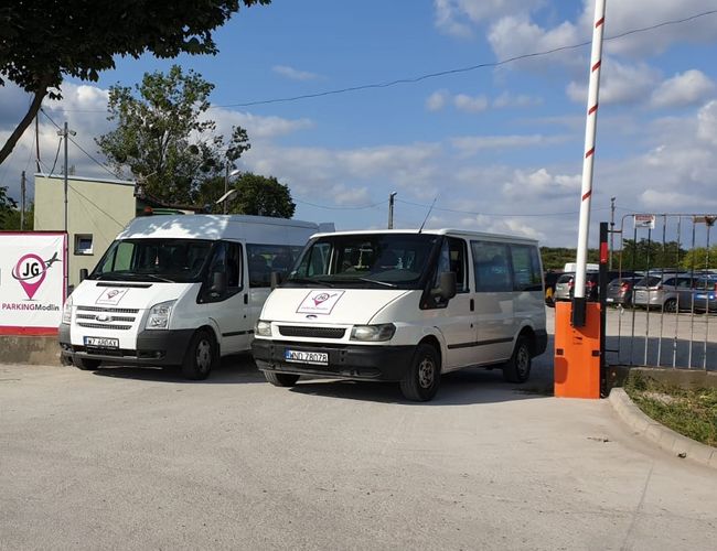 JG Parking - Dos furgonetas blancas están paradas junto a la barrera. El aparcamiento está cercado con una valla metálica. Al fondo se pueden ver coches aparcados.