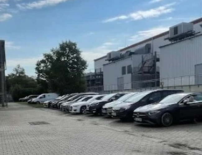 Duży parking z wieloma samochodami stojącymi w rzędach, na tle budynków przemysłowych.