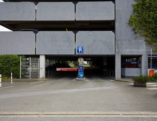 Wjazd do wielopoziomowego parkingu z zadaszonym wjazdem. Nad wjazdem znajduje się betonowa konstrukcja budynku, a po bokach widoczne są elementy zieleni i oznakowanie parkingu.