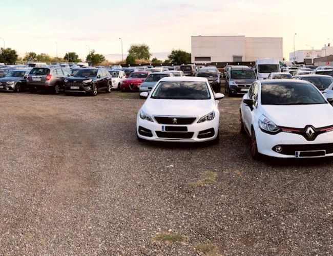 Parking Elite Valet - En el estacionamiento hay coches en una zona de grava. El área es abierta y no está cercada. En el fondo se puede ver un edificio.
