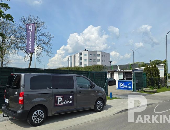 Parkplatz Parkingos - Der schwarze Van steht auf der Einfahrt vor dem Zaun. Im Hintergrund hohe Bäume und Gebäude. Zwei Fahnen mit dem Parkplatz-Logo.
