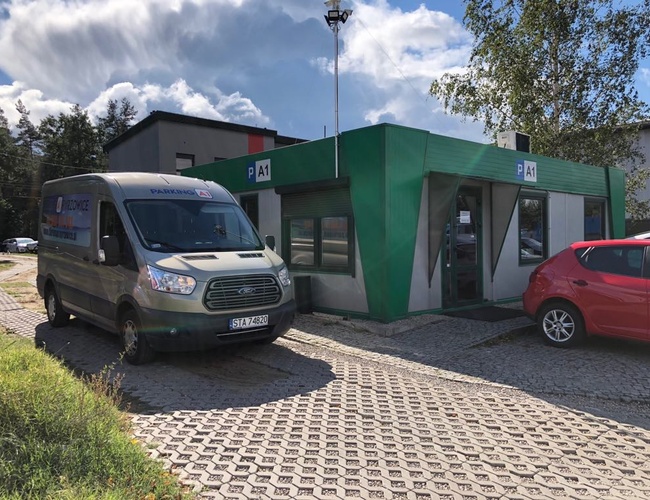 Parking A1 – Na kostce brukowej stoi zielony budynek. Obok zaparkowany jest biały bus i czerwony samochód. W tle widać drzewa i budynki.