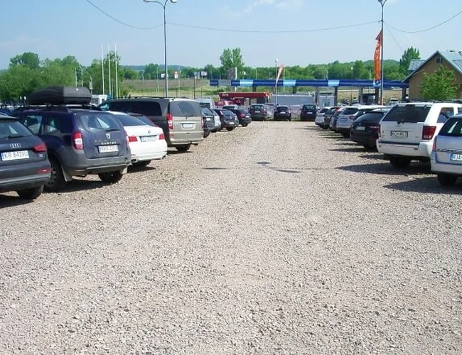 Oranžové parkovisko P3 - štrková plocha s parkujúcimi autami po oboch stranách. Parkovisko nie je ohradené. V pozadí je vidieť zeleň a budovy.