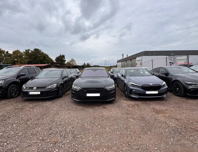 Prime Parking Valet - Pięć samochodów stoi na żwirowym parkingu. W tle widać ogrodzenie z siatki i budynki. Dzień jest pochmurny.