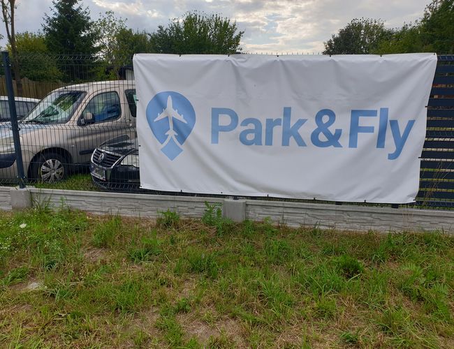 Parking Park&Fly - ogrodzony teren z zaparkowanymi samochodami. Na ogrodzeniu widnieje duży baner.